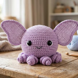 Puede incluir: Un elefante de ganchillo lavanda hecho a mano, con orejas grandes, ojos negros y una boca sonriente. El peluche está sentado sobre una superficie de madera. El elefante tiene pequeños pies rizados. Juguetes de ganchillo.