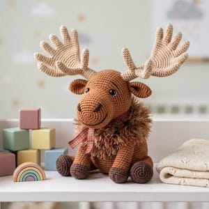 May include: A crocheted brown moose with large antlers, a fluffy neck, and a plaid scarf sits on a white shelf. Beside it are pastel-colored wooden blocks and a rainbow-shaped toy. A folded cream-colored blanket is also on the shelf.