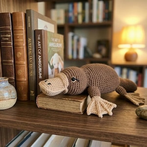 Puede incluir: Un juguete de ornitorrinco de ganchillo marrón hecho a mano con pico y patas beige descansa sobre un libro antiguo. El juguete está junto a un pequeño jarrón con una ramita de lavanda y una colección de libros.