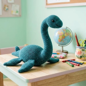 May include: A teal crochet plush toy shaped like a long-necked aquatic dinosaur with a rounded body, four flippers, and a small tail. The creature features a textured yarn surface, a long curved neck, and two small black eyes on its head.
