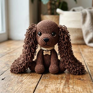 May include: A handmade brown crochet dog with long, curly ears and a small bone-shaped tag that reads "Bailey." The dog is sitting on a wooden surface. The dog's eyes are large and black.