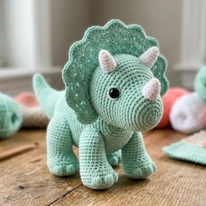 May include: A mint green crocheted triceratops dinosaur toy featuring a large frill, three white horns, and black safety eyes. The stuffed animal is crafted with a textured yarn stitch pattern and stands on four sturdy legs with a pointed tail.