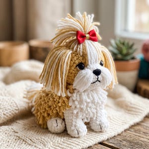 Puede incluir: Un perro Shih Tzu hecho a mano con ganchillo, con una combinación de colores blanco y beige. El perro tiene un lazo rojo y está sentado sobre una manta de punto color crema. El perro tiene ojos y nariz negros.