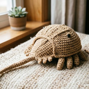 May include: A handmade, crocheted horseshoe crab in shades of tan and brown. The crab has a rounded body, a long tail, and small legs. It rests on a cream-colored, textured surface, with a ball of yarn and a crochet hook nearby.