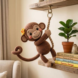 Könnte beinhalten: Ein gehäkelter brauner Affe mit einer gelben Blume im Ohr hängt an einem Seil. Der Affe hat rosige Wangen und lächelt. Der Affe hängt an einem Holzregal mit Büchern und einer kleinen Topfpflanze.
