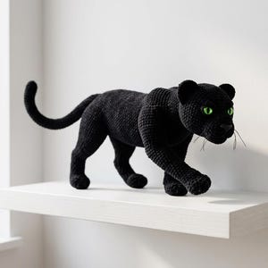 Può includere: Figurina di pantera nera fatta a mano con occhi verde brillante. La pantera è all'uncinetto e posa in posizione di camminata. La figurina è esposta su una mensola bianca.