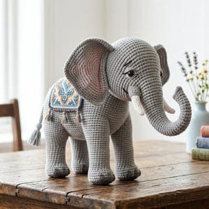 Könnte beinhalten: Ein grauer gehäkelter Elefant mit großen Ohren, einem gerollten Rüssel und weißen Stoßzähnen. Er hat einen dekorativen Sattel mit blauen und orangefarbenen Mustern und Quasten. Der Elefant steht auf einer Holzoberfläche.