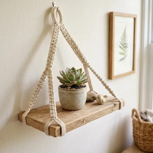 May include: A rectangular wooden shelf suspended by cream-colored macrame cord. The shelf holds a small speckled ceramic pot containing a green succulent plant and a small stone bird figurine. The macrame features a knotted design with a top loop.