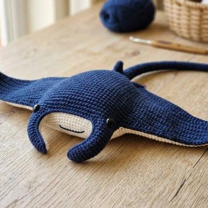May include: A handmade crochet manta ray toy featuring a deep navy blue top and a cream-colored underside. The plush creature has two small black plastic eyes and a simple stitched mouth, with wide fins and a long, slender tail extending behind it.