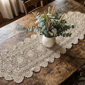 Pode incluir: Um caminho de mesa de crochê bege com um padrão de renda intrincado e bordas festonadas. Um vaso de cerâmica branca fica no centro, preenchido com um buquê de folhas de eucalipto verdes, pequenas flores brancas e hastes de trigo secas.