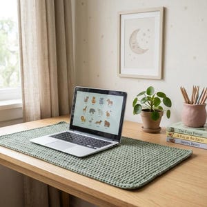 Op de afbeelding: Een rechthoekige saliegroene gehaakte bureauonderlegger met een gestructureerd steekpatroon, geplaatst op een houten oppervlak onder een zilveren laptop. De mat heeft een uniform geweven ontwerp met afgeronde hoeken.