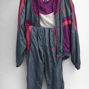 May include: A vintage Nike tracksuit in a gray, purple, and pink color scheme. The jacket has a hood and pink stripes, and the pants are a matching gray. The set is displayed on a hanger against a white background.