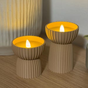 May include: Two beige candle holders with a ribbed design, each holding a glowing yellow candle. The candles have a realistic flame effect. The holders are on a wooden surface, with a decorative vase in the background.