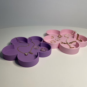 Puede incluir: Dos bandejas de joyería en forma de flor, una lavanda y otra rosa, están una al lado de la otra. La bandeja lavanda contiene un collar y anillos dorados. La bandeja rosa exhibe anillos, collares y pulseras. Estas bandejas son ideales para organizar y exhibir joyas.