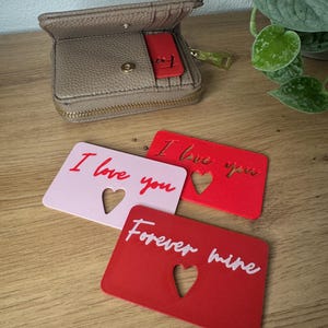 May include: A taupe leather wallet with a gold zipper and a red tag. Three small cards with heart cutouts and the phrases "I love you" and "Forever mine" are displayed on a wooden surface.