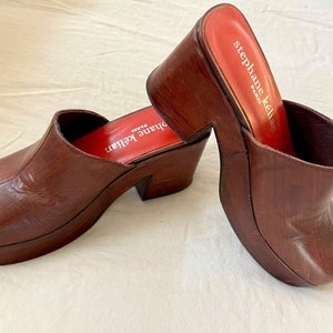 May include: A pair of brown leather platform clogs with a red label that reads "Stephane Kelian Paris".