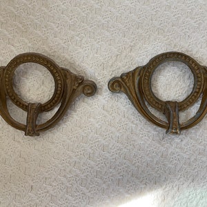 May include: Two antique gold-toned metal drawer pulls with ornate designs. Each pull features a circular ring with a beaded border and a decorative scroll design on either side.