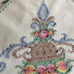 May include: White linen tablecloth with embroidered floral design. The design features a brown vase with blue and green accents, filled with pink, red, yellow, and orange flowers.