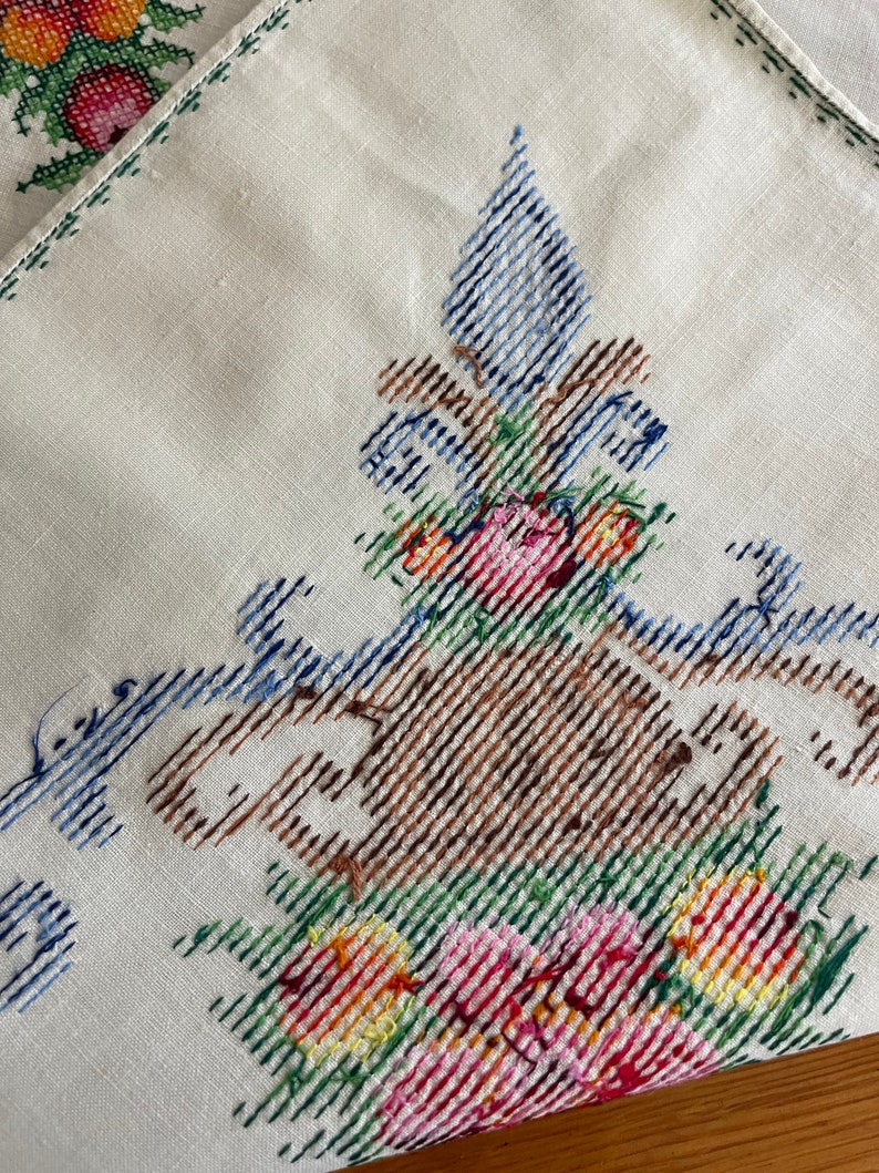 May include: White linen tablecloth with embroidered floral design. The design features a brown vase with blue and green accents, filled with pink, red, yellow, and orange flowers.
