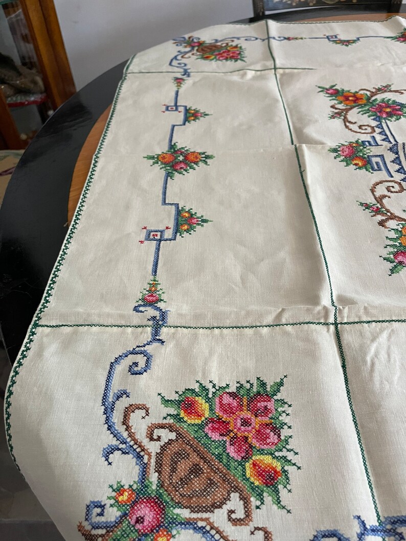 May include: A white linen tablecloth with a green border and embroidered floral designs in shades of red, pink, yellow, blue, and green.