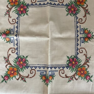 May include: A white linen tablecloth with a hand-stitched embroidered border. The border features a repeating pattern of stylized flowers in shades of orange, red, green, and blue.