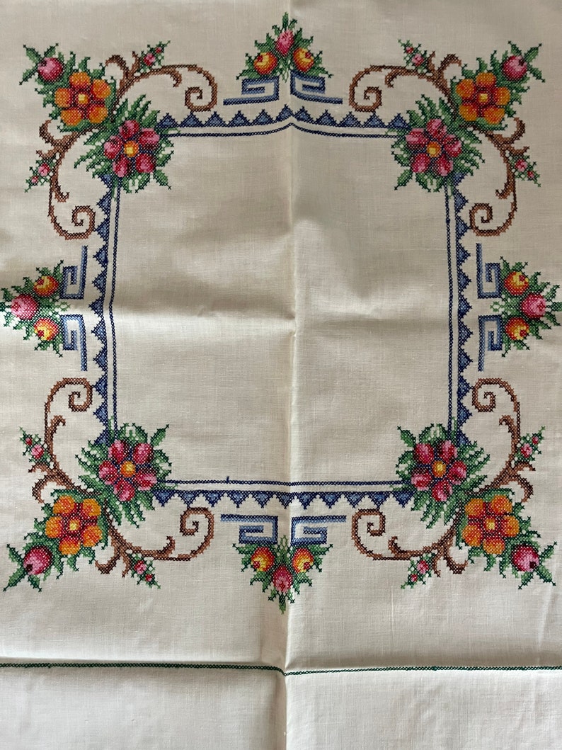 May include: A white linen tablecloth with a hand-stitched embroidered border. The border features a repeating pattern of stylized flowers in shades of orange, red, green, and blue.
