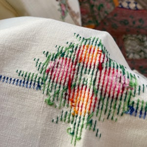 May include: Close-up of a white fabric with a colorful cross-stitch design. The design features green, red, orange, yellow, and pink threads.