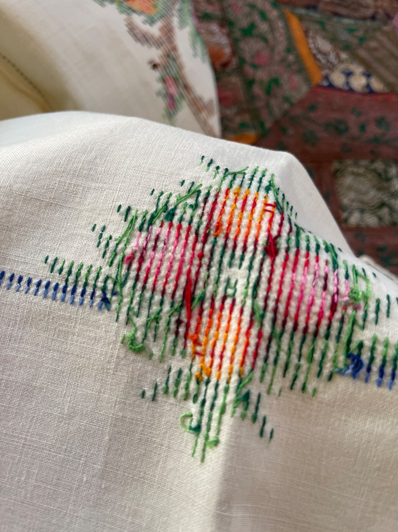 May include: Close-up of a white fabric with a colorful cross-stitch design. The design features green, red, orange, yellow, and pink threads.