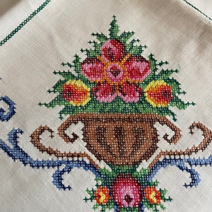 May include: White linen tablecloth with a cross-stitch design of a vase with flowers. The vase is brown and the flowers are pink, red, yellow, and green.