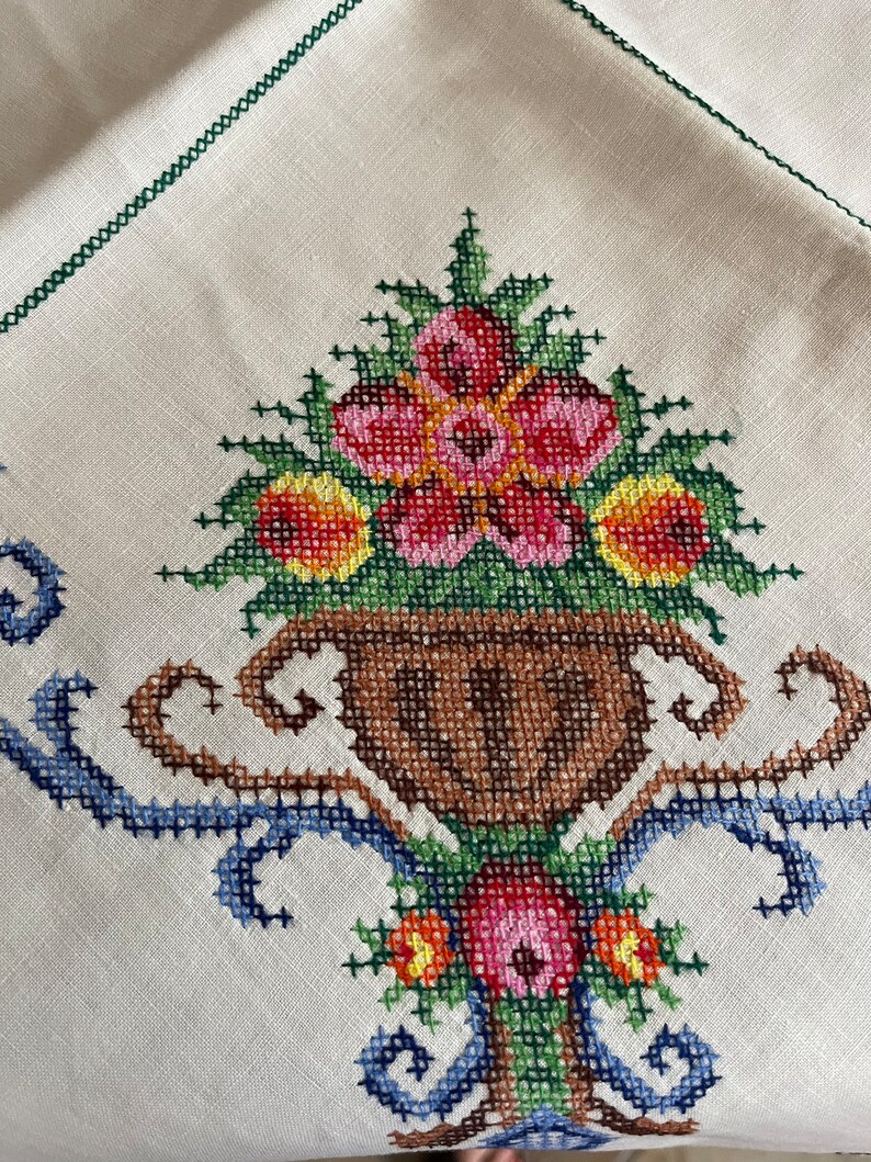 May include: White linen tablecloth with a cross-stitch design of a vase with flowers. The vase is brown and the flowers are pink, red, yellow, and green.