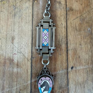 May include: A silver-toned metal watch fob chain featuring a rectangular central link with a pink and blue enamel geometric pattern. A swivel connector attaches to a horseshoe-shaped pendant displaying a black horse silhouette on a pink background.