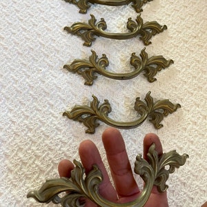 May include: Five ornate gold-toned cabinet drawer pulls with a decorative scroll design. The pulls are arranged in a row, with the bottom pull being held in a hand.