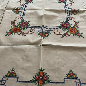 May include: White linen tablecloth with a floral embroidered border. The border features a repeating pattern of stylized flowers in shades of red, orange, yellow, and green. The tablecloth has a simple, elegant design.