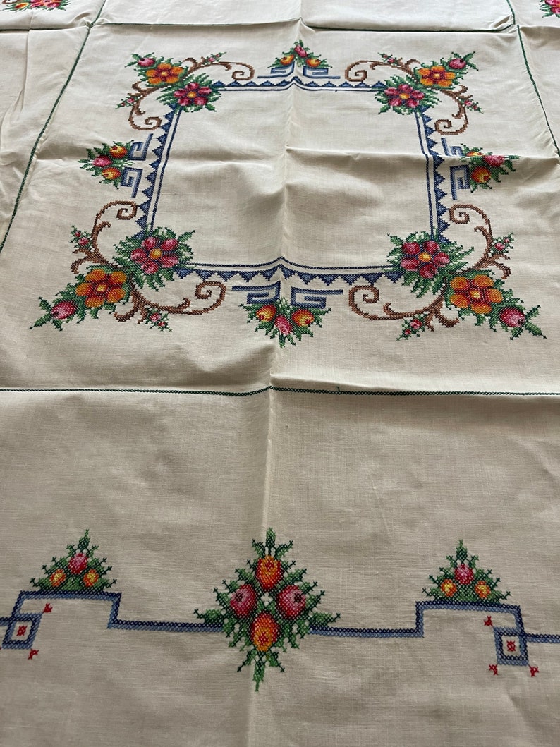 May include: White linen tablecloth with a floral embroidered border. The border features a repeating pattern of stylized flowers in shades of red, orange, yellow, and green. The tablecloth has a simple, elegant design.