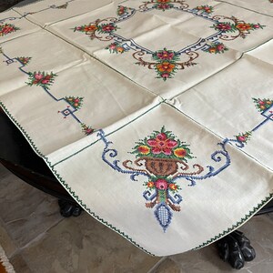 May include: A white linen tablecloth with a green border and embroidered floral designs in shades of pink, orange, yellow, blue, and green.