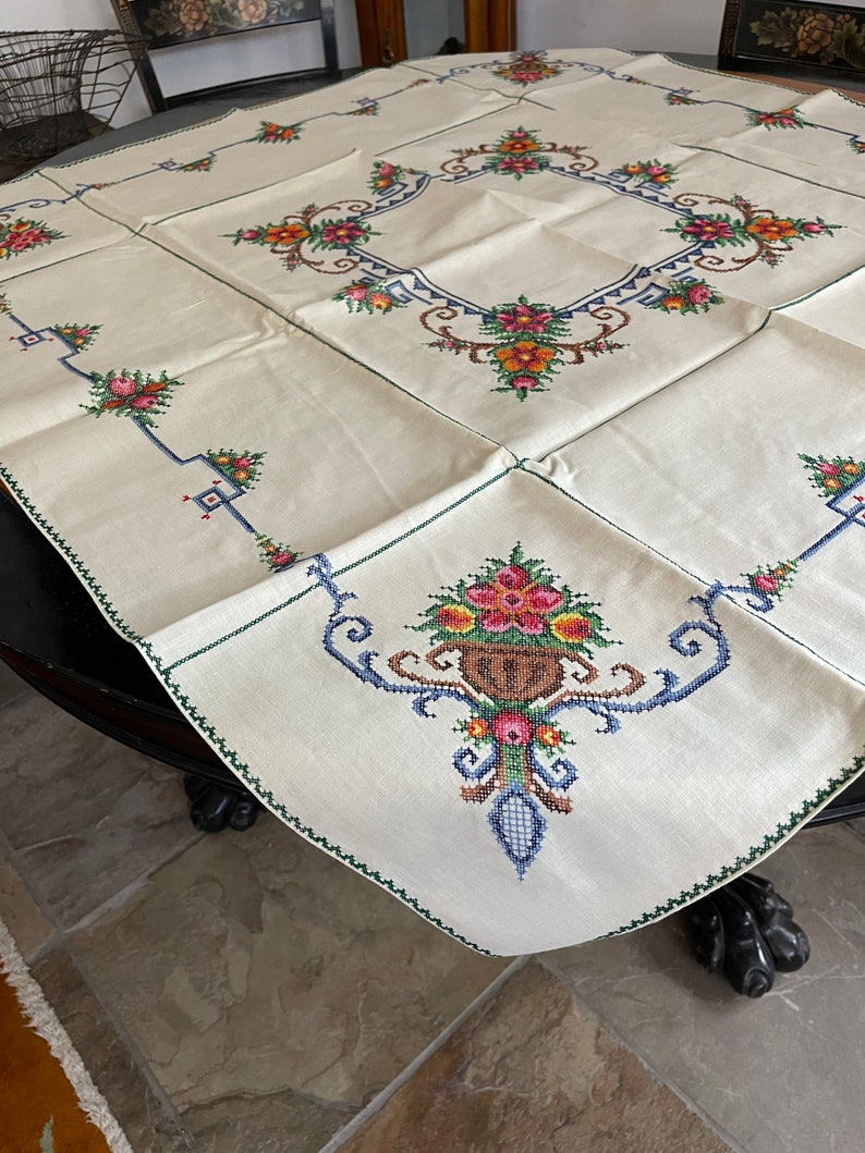 May include: A white linen tablecloth with a green border and embroidered floral designs in shades of pink, orange, yellow, blue, and green.
