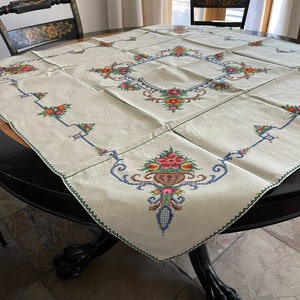 May include: A white linen tablecloth with embroidered floral designs in shades of pink, yellow, blue, and green. The tablecloth is laid on a round wooden table with a black finish.