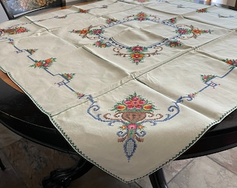 May include: A white linen tablecloth with embroidered floral designs in shades of pink, yellow, blue, and green. The tablecloth is laid on a round wooden table with a black finish.