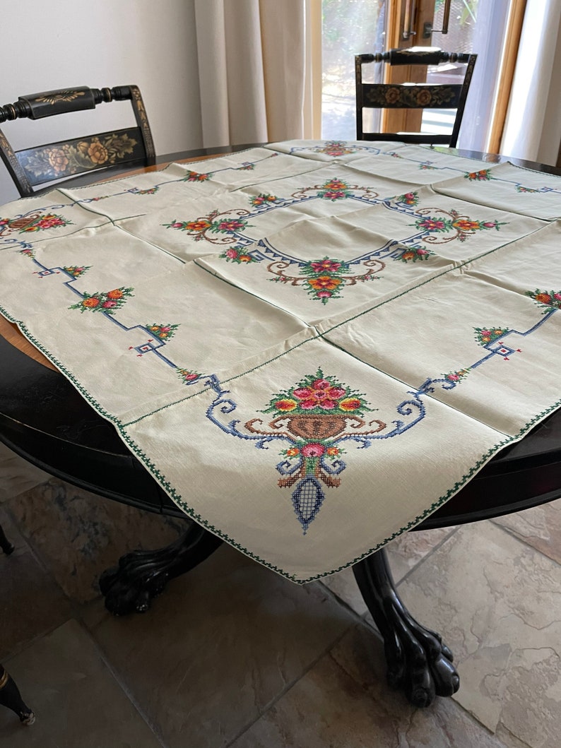 May include: A white linen tablecloth with embroidered floral designs in shades of pink, yellow, blue, and green. The tablecloth is laid on a round wooden table with a black finish.