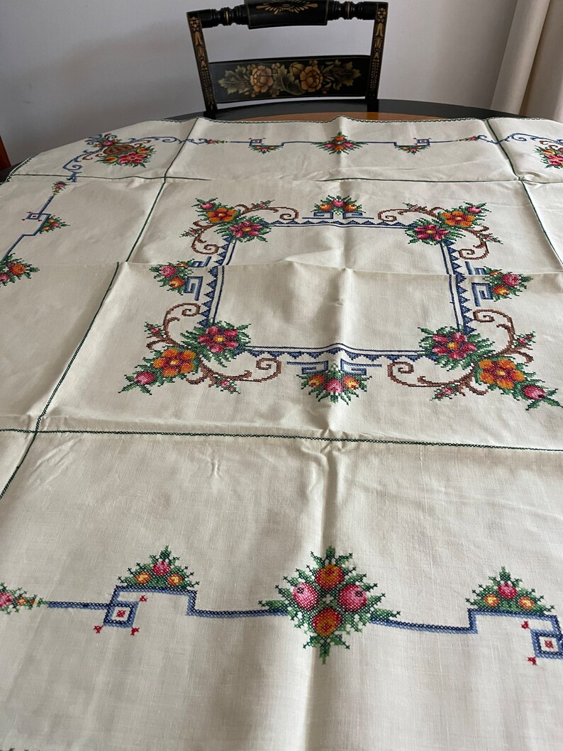 May include: White linen tablecloth with a green, blue, and orange embroidered floral border.