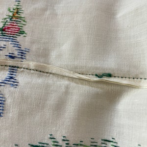 May include: A white linen tablecloth with a green, red, and yellow embroidered floral design. The tablecloth has a seam down the center.