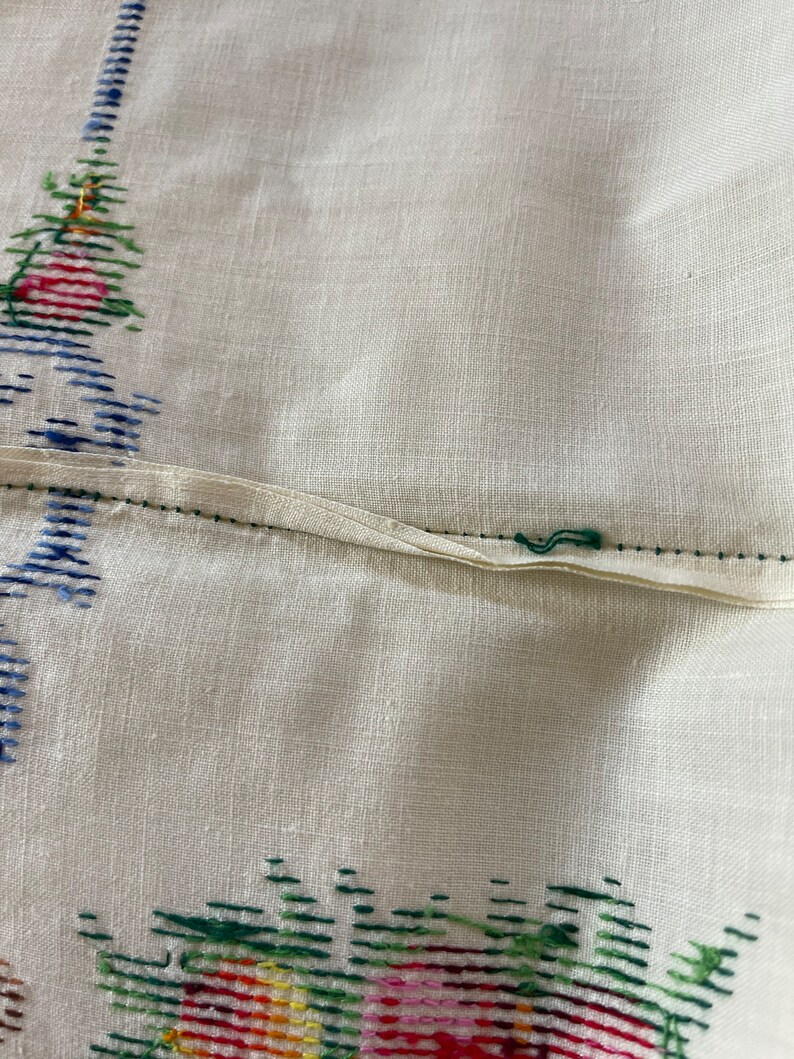 May include: A white linen tablecloth with a green, red, and yellow embroidered floral design. The tablecloth has a seam down the center.