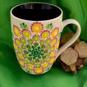 Puede incluir: Una taza de cerámica blanca con interior negro y un colorido diseño de mandala con puntos amarillos, verdes y rosas. La taza está sobre una tela verde junto a un grupo de cristales de color claro.