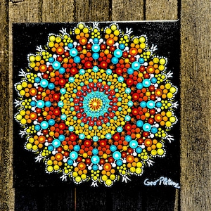 Puede incluir: Una colorida pintura de mandala sobre un lienzo cuadrado negro. El diseño presenta un patrón simétrico de puntos vibrantes en tonos de amarillo, naranja, rojo y turquesa. La obra está firmada en la esquina inferior derecha.