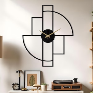 May include: A contemporary wall clock featuring a black geometric design with a circular black clock face and gold hands. The design includes rectangular and semi-circular shapes. The clock is mounted on a white wall. The background shows a lamp and shelves.