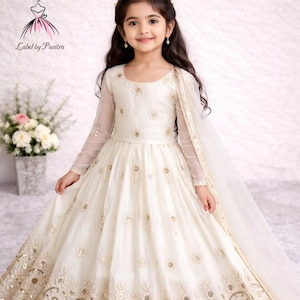 Puede incluir: Un vestido crema para niña con mangas largas y transparentes y bordados dorados intrincados. El vestido presenta una falda amplia y un chal transparente a juego. El texto "LABEL BY PAVITRA" es visible en la parte inferior del vestido.