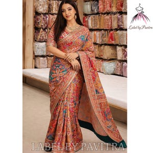 Puede incluir: Una mujer con un sari vibrante con intrincados diseños florales y de cachemira en tonos rojos, naranjas y turquesas. El sari tiene un borde detallado y está etiquetado como "Label by Pavitra".