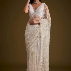 May include: A white saree featuring intricate silver embroidery and sequin detailing throughout the sheer fabric. The garment includes a sleeveless blouse with a deep V-neckline and a matching border with scalloped edges along the draped pallu.