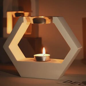 Könnte beinhalten: Ein weißer, geometrischer Kerzenhalter mit sechseckigem Design. Eine brennende Teelichtkerze befindet sich in der Mitte, mit einer weiteren Teelichtkerze darüber. Der Halter hat eine moderne, minimalistische Ästhetik mit warmer Beleuchtung.