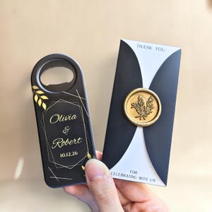 Pode incluir: Um abridor de garrafas preto com linhas geométricas douradas e o texto Olivia & Robert 10,12,26, acompanhado de um cartão preto e branco com o texto THANK YOU e FOR CELEBRATING WITH US, decorado com um selo de cera dourado com folhas.